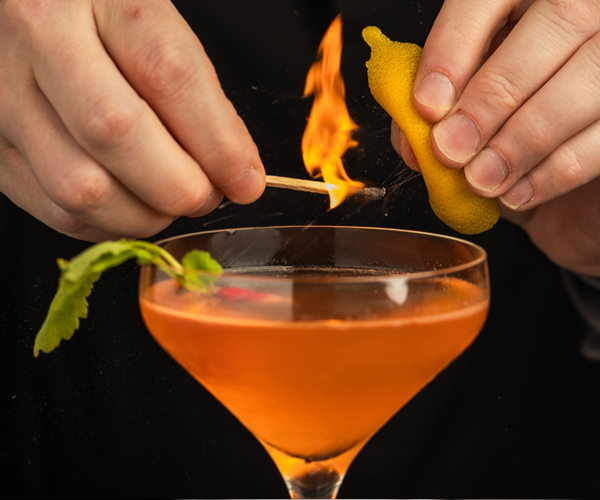 A bartender ignites a cocktail with a match while holding a lemon peel over the glass.
