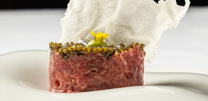 A round piece of steak tartare topped with caviar, a yellow edible flower, and a decorative white crisp served on a white plate.