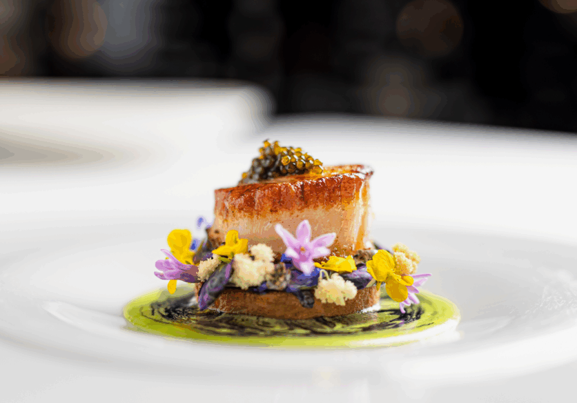 A seared scallop topped with caviar, served on a round base, garnished with edible flowers, herbs, and colorful sauce on a white plate.