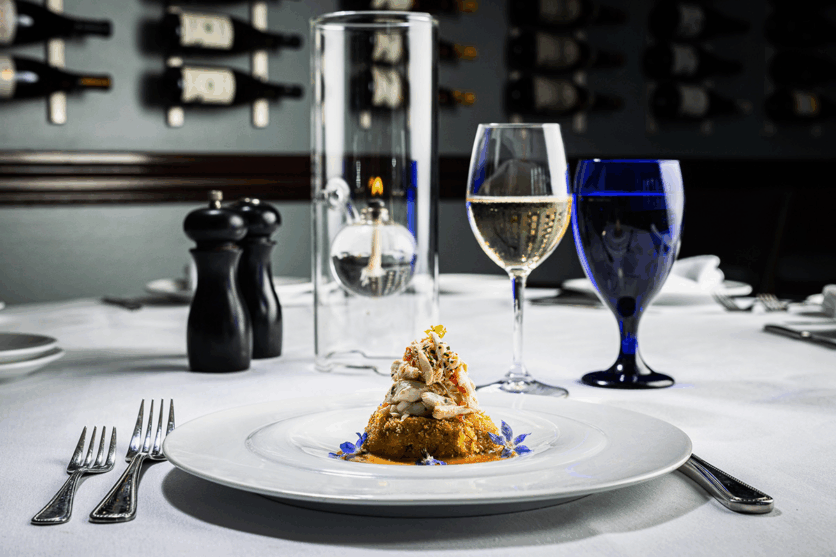 A plated gourmet dish is set on a white tablecloth, accompanied by a glass of white wine, a blue water glass, a pepper grinder, and a candle, with wine bottles in the background.