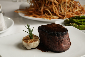 A thick, dark steak sits on a white plate next to a roasted garlic bulb with a sprig of rosemary.
