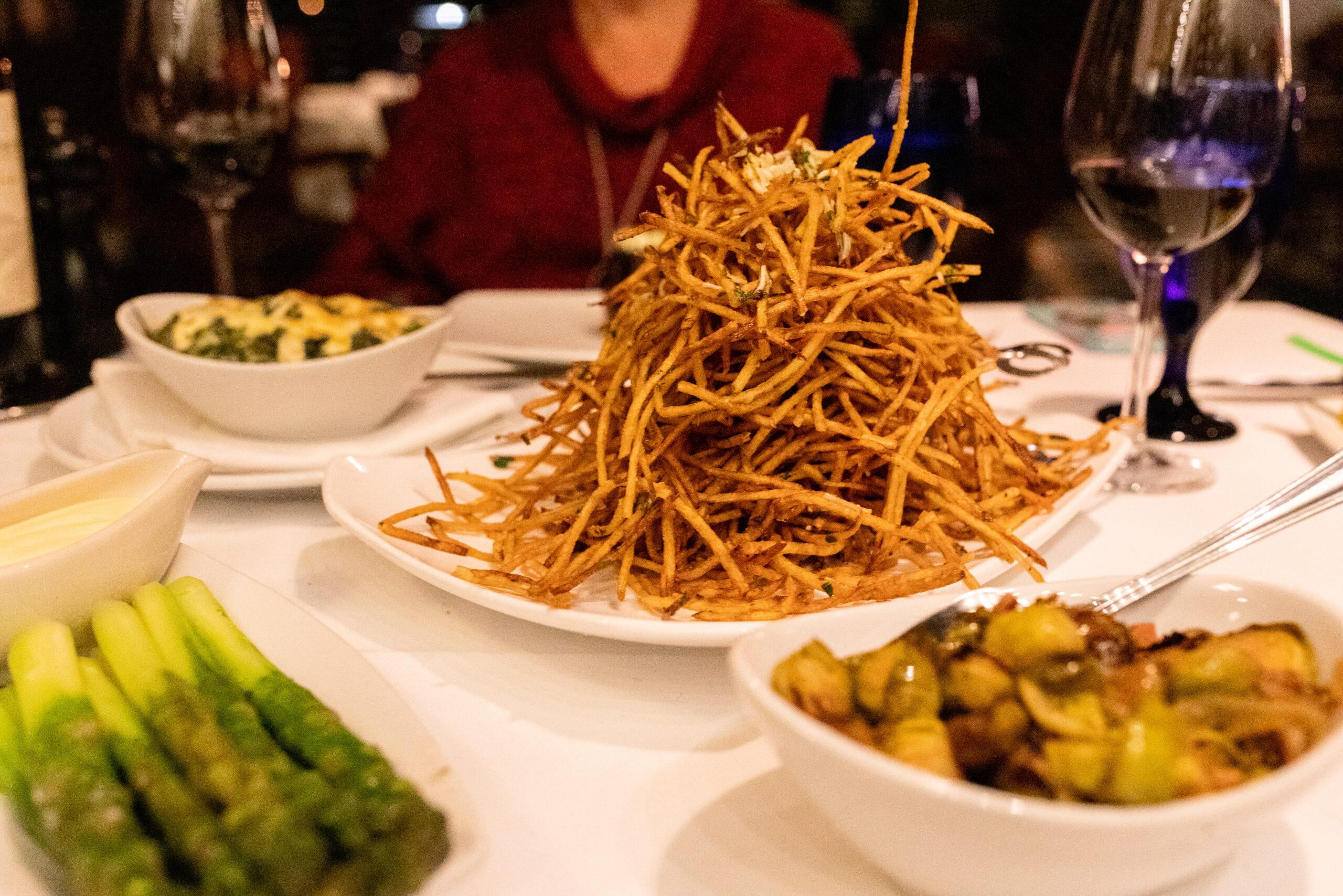 A large pile of crispy shoestring fries sits on a white plate, with bowls of asparagus, Brussels sprouts, and a creamy dip surrounding it.