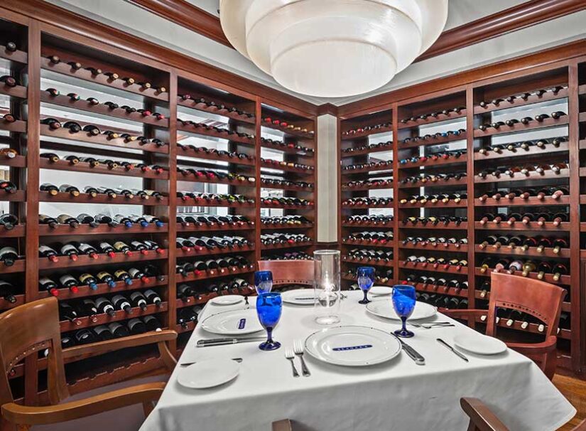 A dining table set for four with blue water glasses, surrounded by wooden wine racks filled with bottles in a wine cellar.