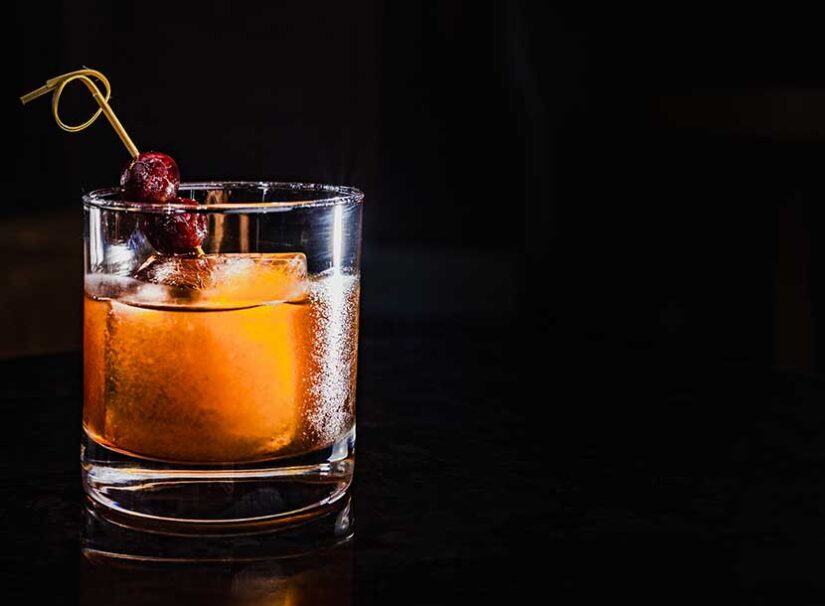 A glass of amber cocktail with a large ice cube and two cherries on a cocktail pick, set against a dark background.