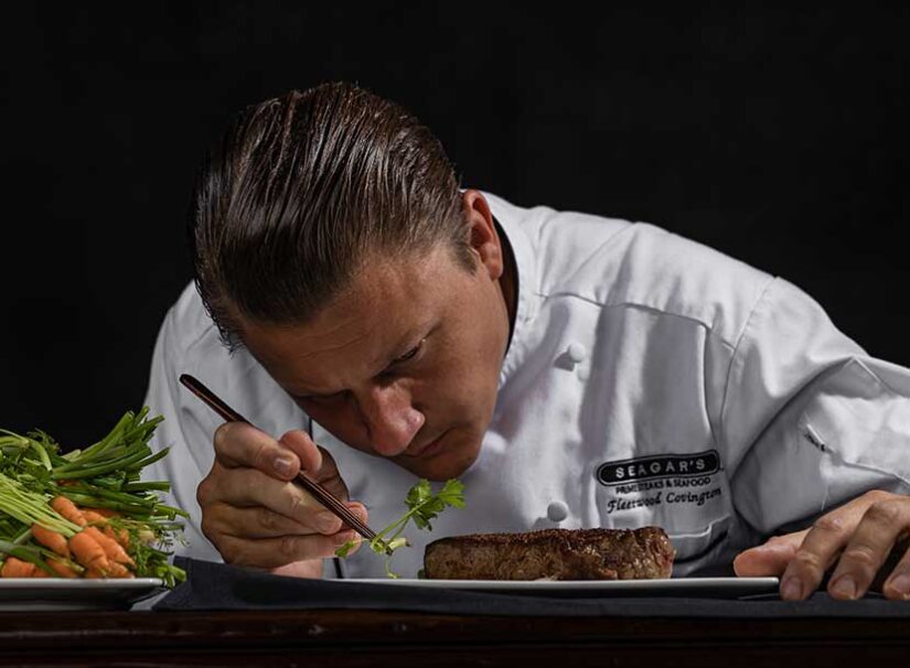 A chef in a white uniform uses tweezers to place herbs on a cooked steak, with carrots and greens nearby on a dark background.