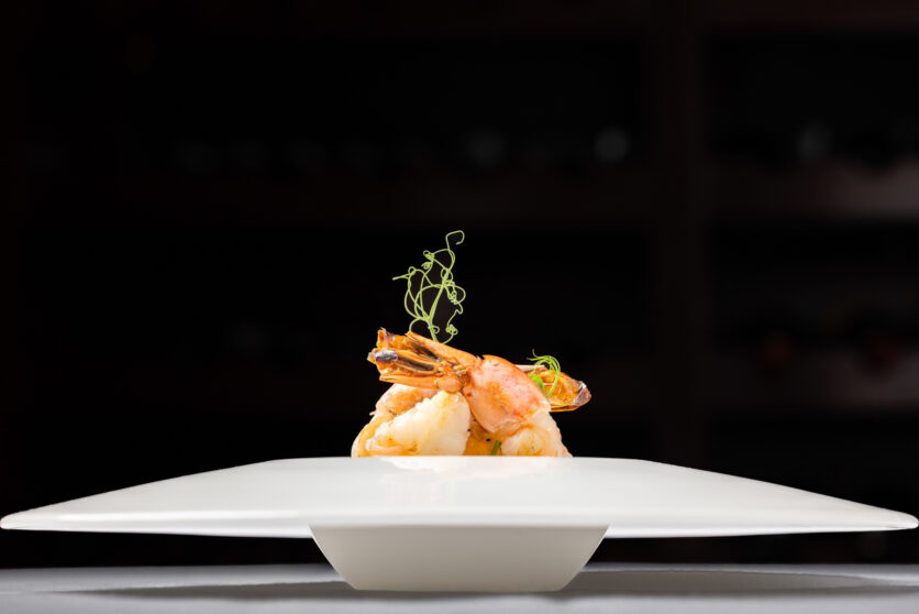 A gourmet dish featuring two cooked shrimp, garnished with microgreens, is arranged on a white plate against a dark background.
