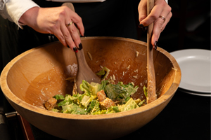 A person tossing a Caesar salad in a wooden bowl with two wooden salad servers.