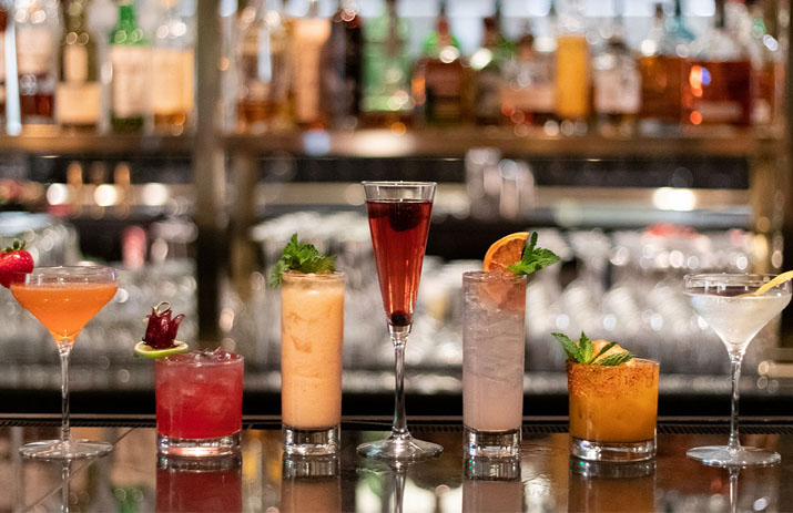 A row of six different colorful cocktails is displayed on a bar counter, each garnished with fruit or herbs, with liquor bottles blurred in the background.