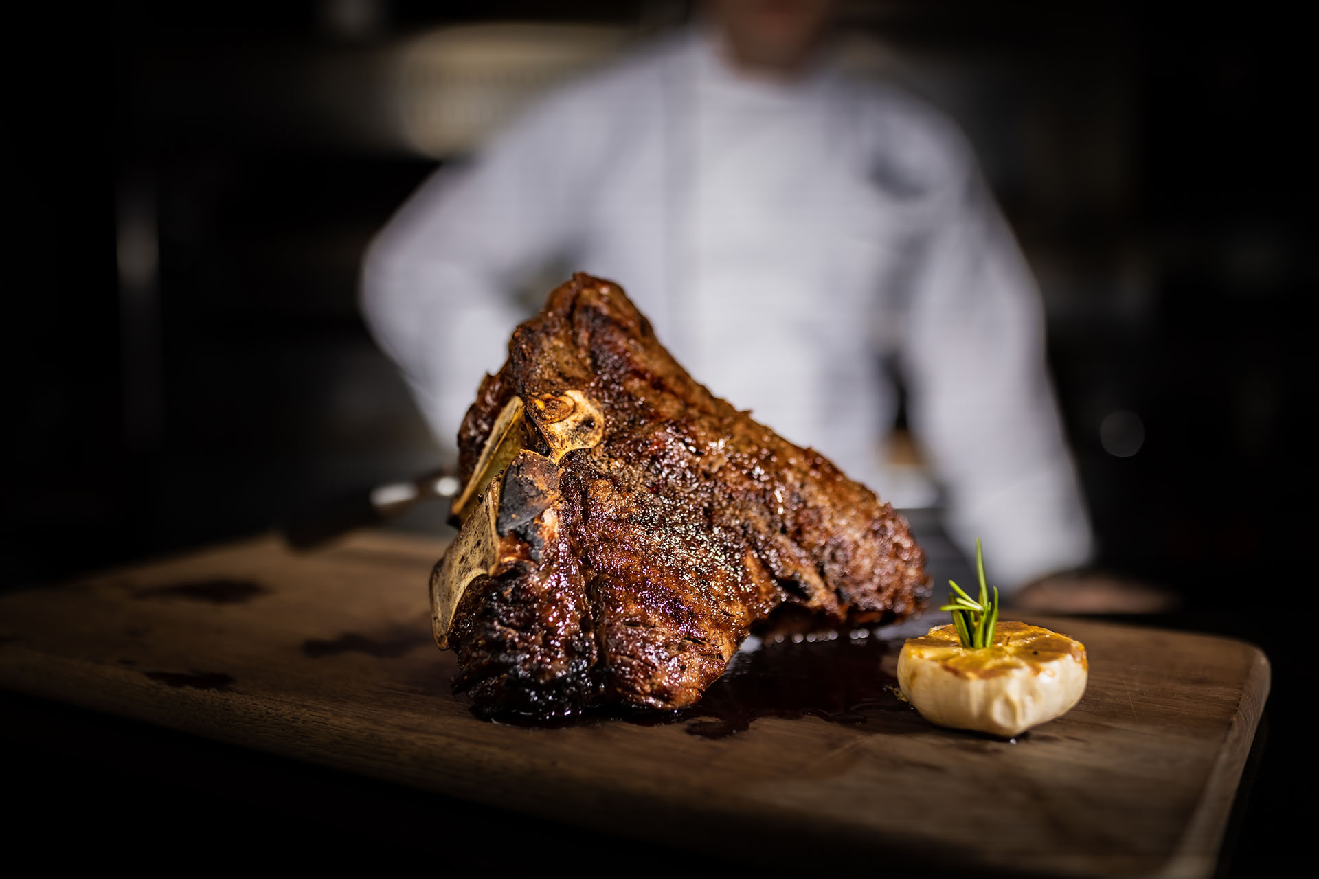 A large, cooked T-bone steak sits on a wooden board beside a halved roasted garlic, with a chef in a white uniform blurred in the background.