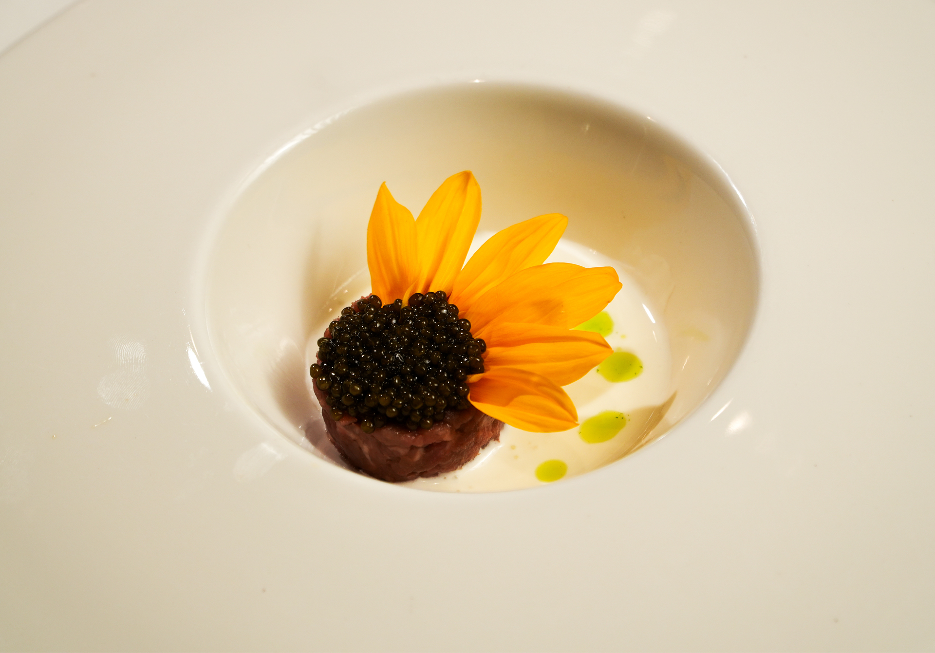 A round dish with a small portion of food topped with black caviar, a few bright yellow flower petals, white sauce, and green oil drops, presented in a white bowl.
