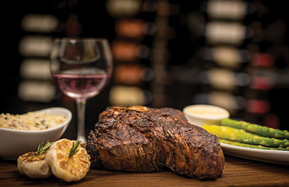 A grilled steak with a side of asparagus, roasted garlic, a dish of rice, a small bowl of sauce, and a glass of red wine on a wooden table.