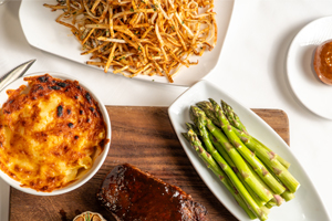 A meal with grilled asparagus, baked macaroni and cheese, shredded hash browns, and a glazed meatloaf on a wooden board, viewed from above.