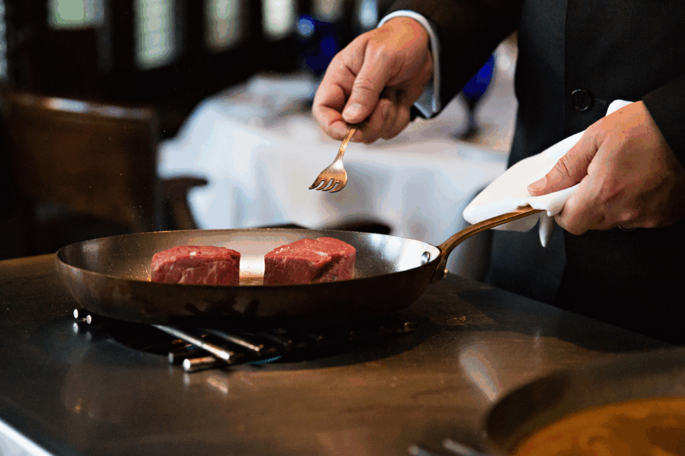 A person in formal attire sears two pieces of raw steak in a frying pan on a stovetop, holding a small fork and a towel.