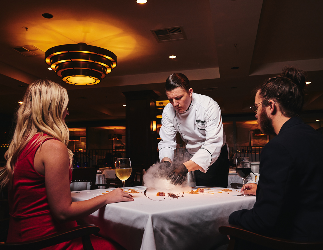 A chef prepares a dish with smoke at a table where a couple dines in a restaurant.