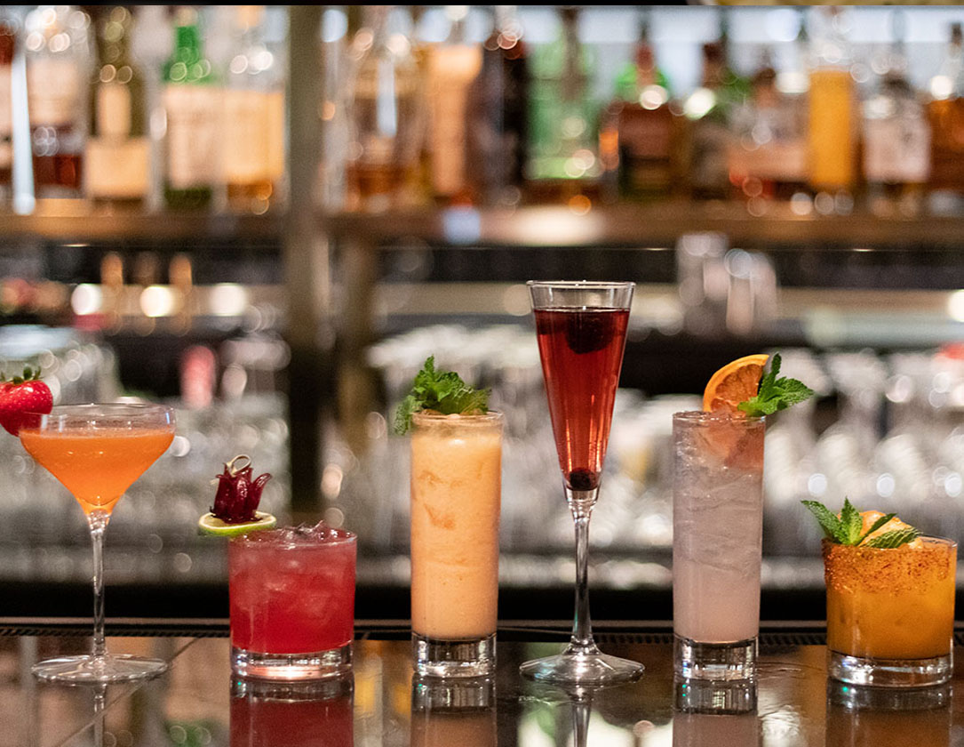 A row of six colorful cocktails garnished with fruit and mint sits on a bar counter in front of a blurred background of bottles.