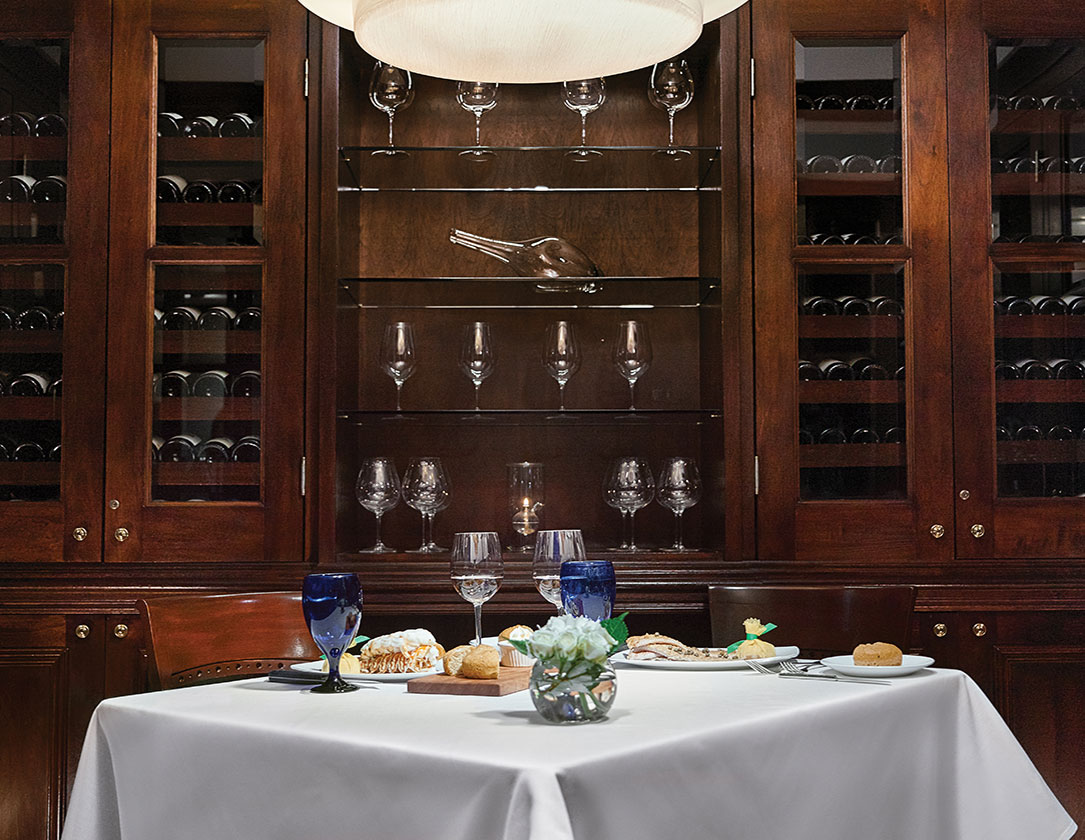 A white tablecloth covers a dining table set with plates, glasses, and a floral arrangement in front of a dark wood wine cabinet.