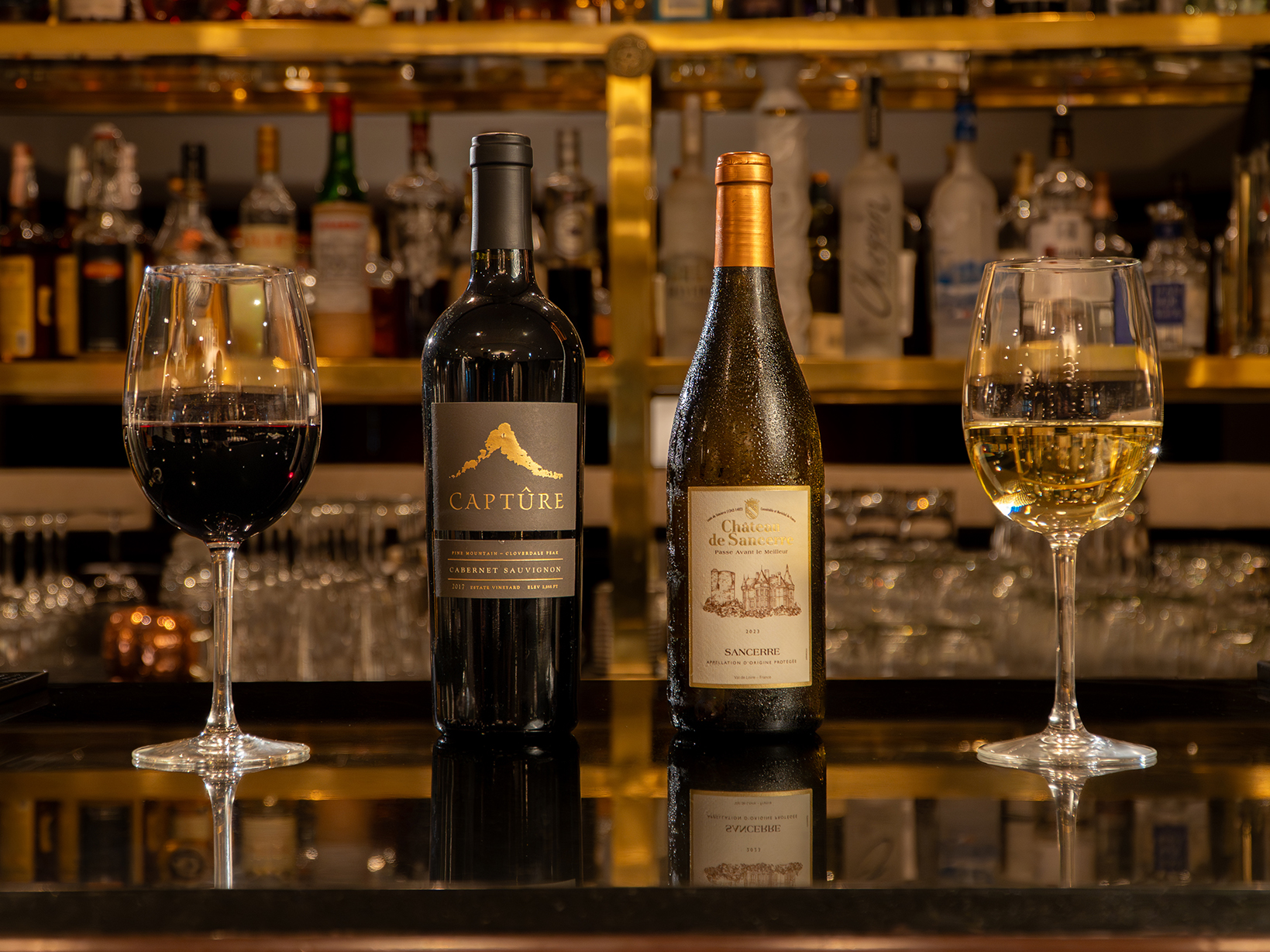 Two bottles of wine, one red and one white, are displayed on a bar with matching filled wine glasses and various liquor bottles in the background.