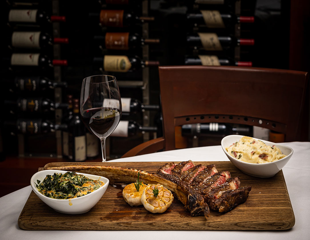 A sliced tomahawk steak is served with creamed spinach, garlic, and a side of pasta on a wooden board, with a glass of red wine and a wine rack in the background.