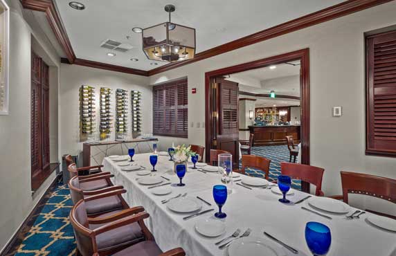A formal dining room set with a long table, white tablecloth, blue glasses, and wooden chairs; wine rack and bar area visible in the background.