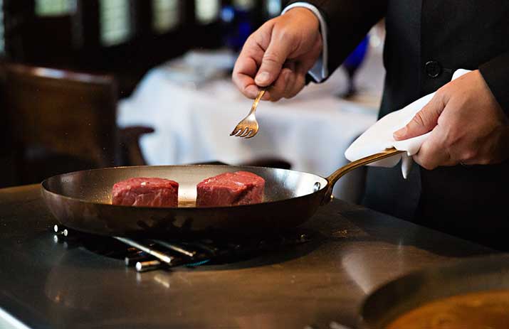 A person in formal attire cooks two raw steaks in a frying pan on a stovetop, holding a fork in one hand and a napkin in the other.