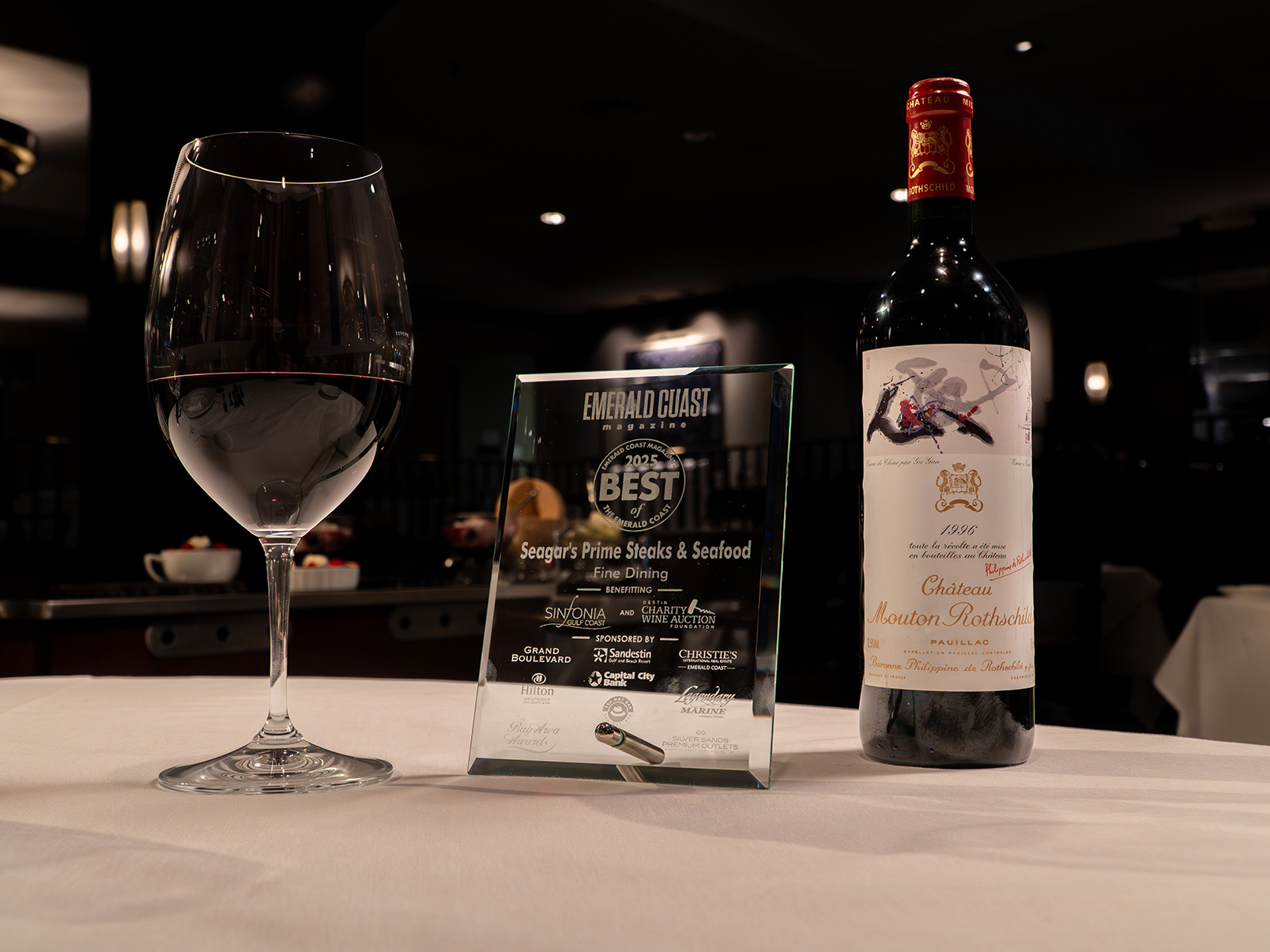 A glass of red wine, a bottle of Château Mouton Rothschild wine, and an award plaque sit on a white tablecloth in a dimly lit restaurant.