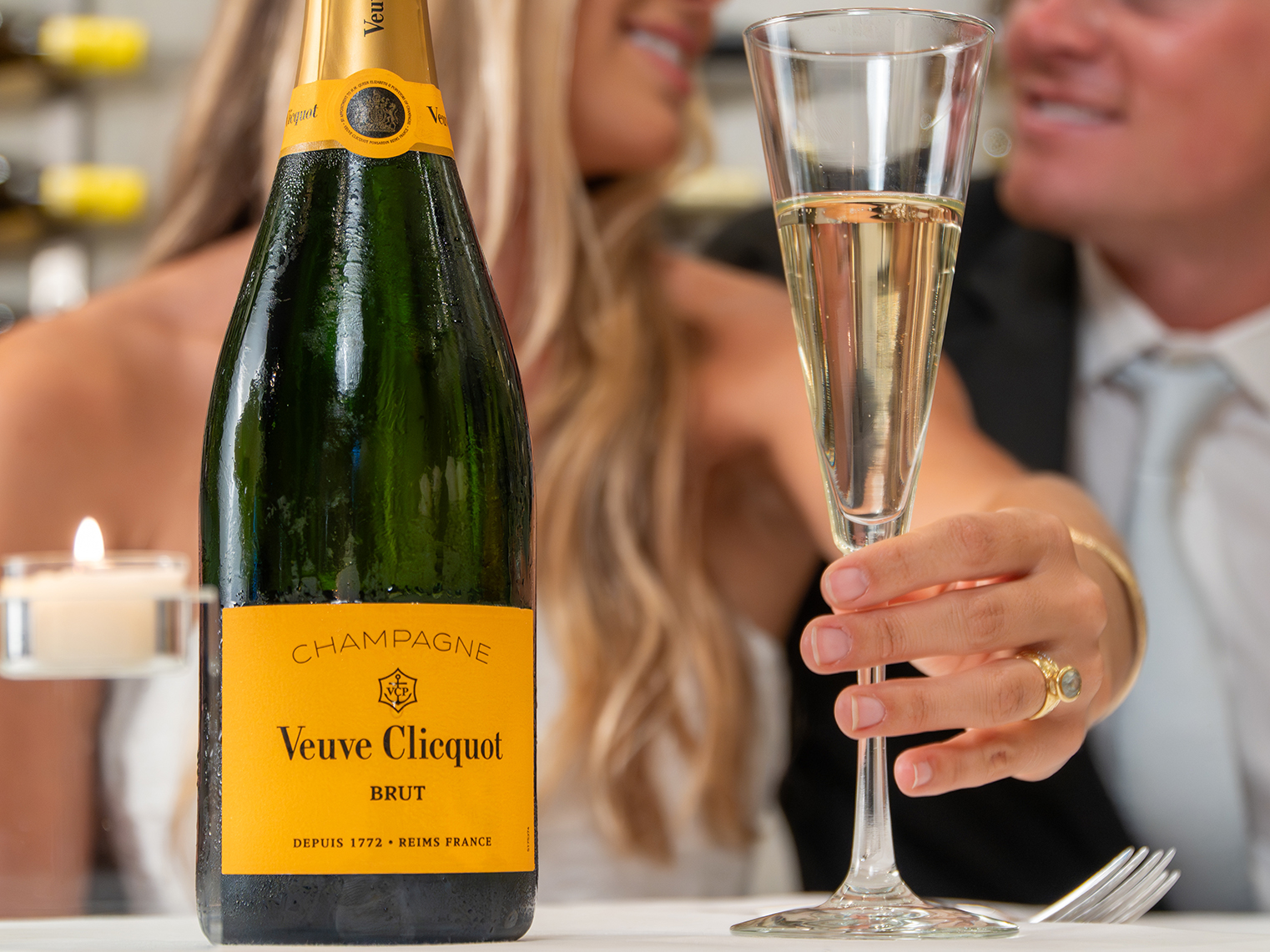 A bottle of Veuve Clicquot Brut champagne and a filled champagne flute on a table, with two people in formal attire in the background.