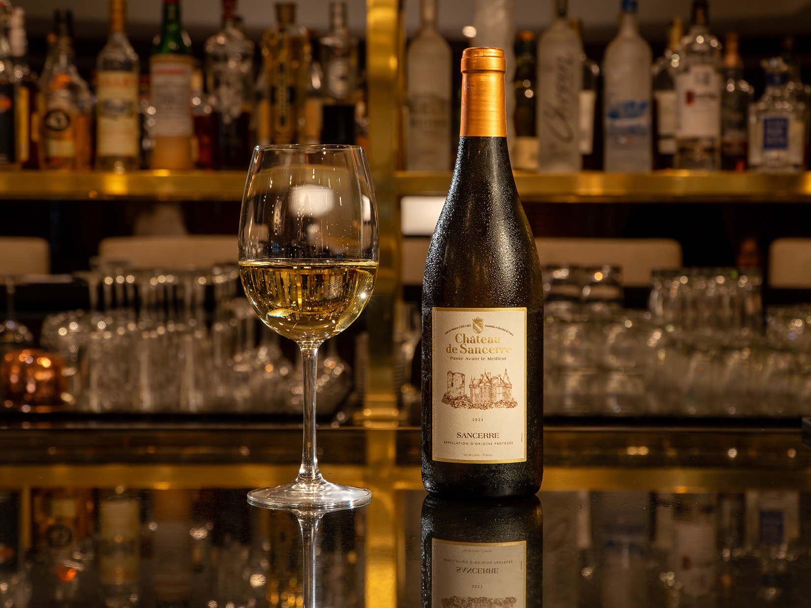 A bottle of Château de Sancerre white wine and a filled wine glass are placed on a bar counter with bottles and glasses in the background.