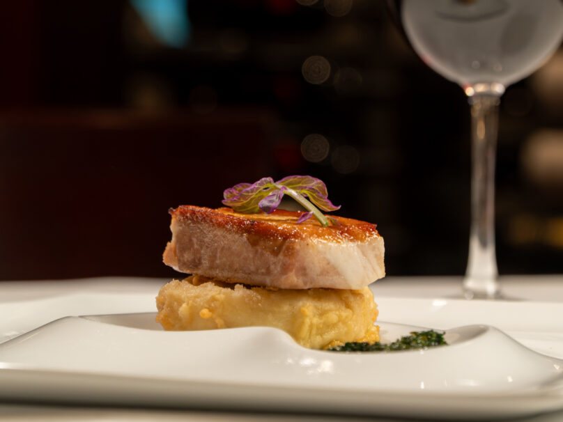 A seared piece of fish is plated on top of a golden, battered base and garnished with microgreens, served on a white plate with a wine glass in the background.