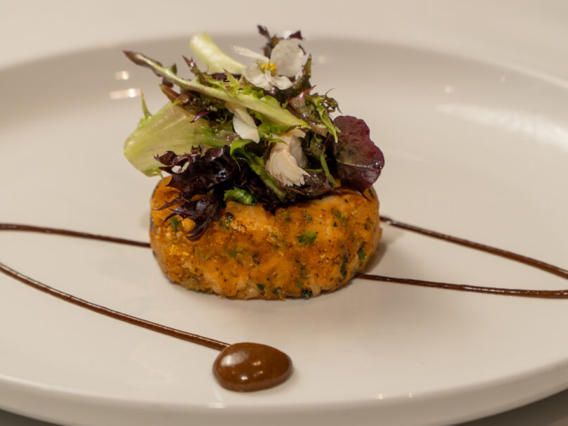 A round patty topped with mixed greens and edible flowers is plated with two lines and a drop of brown sauce on a white dish.