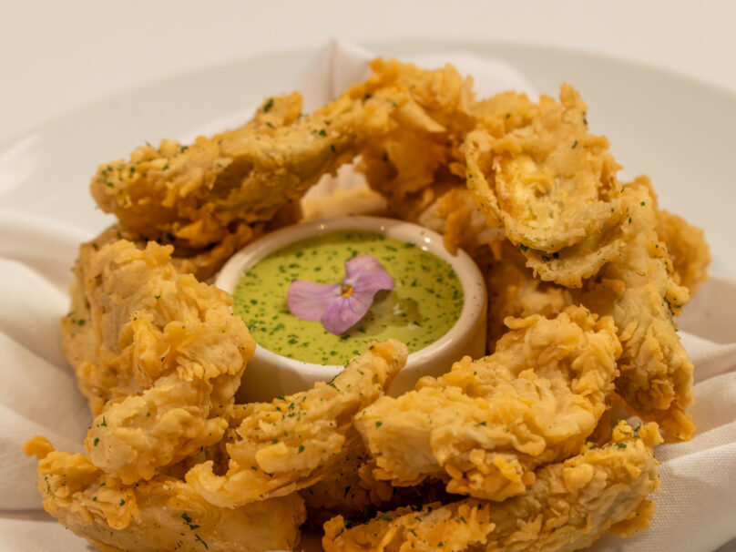A plate of fried oyster mushrooms arranged around a small bowl of green dipping sauce, garnished with a purple edible flower.