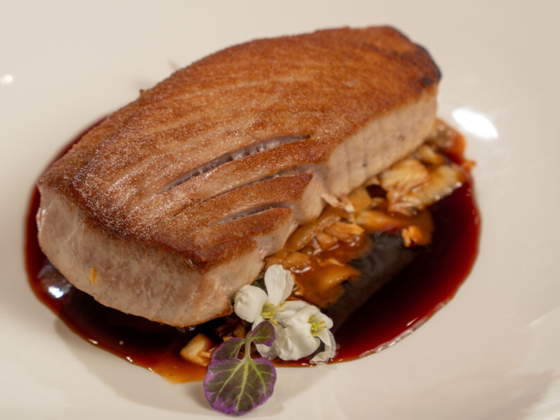 A seared piece of fish sits on a dark sauce with chopped mushrooms, garnished with a small edible flower and green leaf.