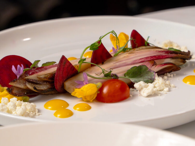 A plated gourmet salad with endive, beet slices, cherry tomato, edible flowers, microgreens, crumbled cheese, and yellow sauce dots on a white dish.