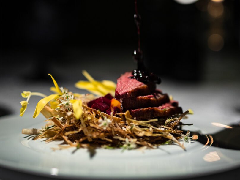 A gourmet dish featuring sliced meat atop a bed of crispy shredded vegetables, garnished with edible flowers, with sauce being poured over the top.
