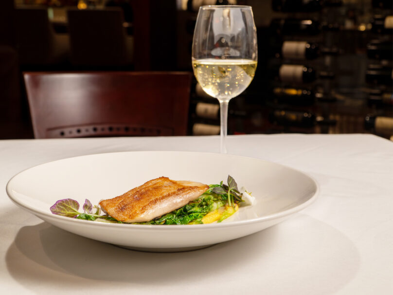 A plated fish fillet with greens and garnish is served on a white tablecloth beside a glass of white wine in a restaurant setting.