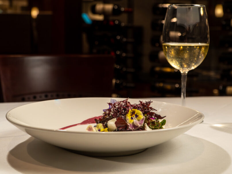 A plated gourmet salad with edible flowers and a glass of white wine on a restaurant table, with a wine rack in the background.