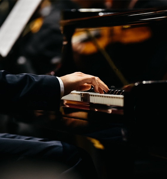 A person in formal attire plays a grand piano, with string musicians and sheet music visible in the blurred background.