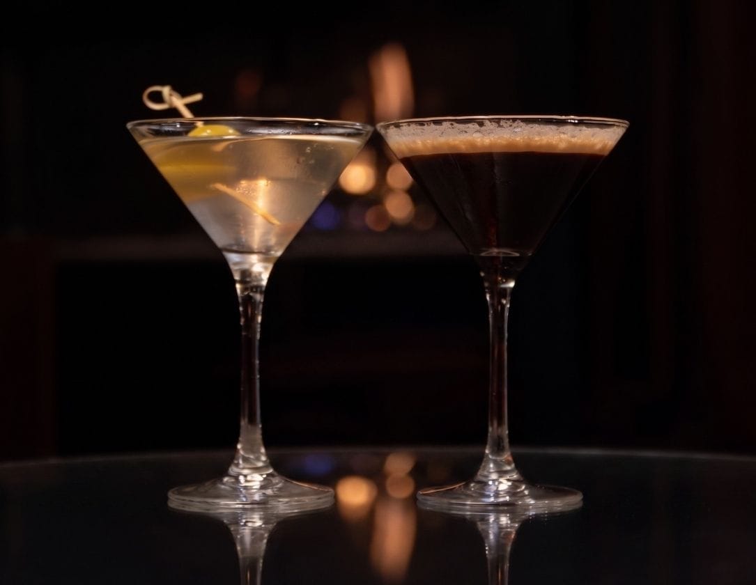 Two martini glasses on a glass table, one with a clear cocktail and lemon twist garnish, the other with a dark cocktail and a light frothy top, against a dark, blurred background.