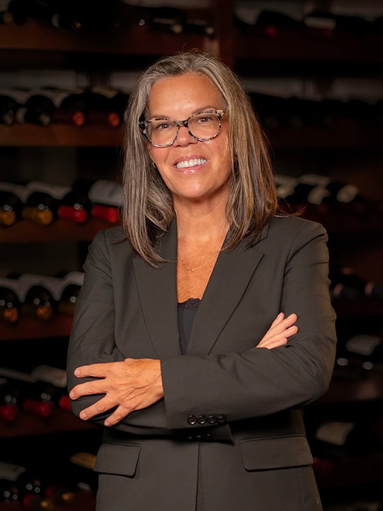 A woman with gray hair and glasses, wearing a dark blazer, stands with arms crossed in front of shelves filled with wine bottles.